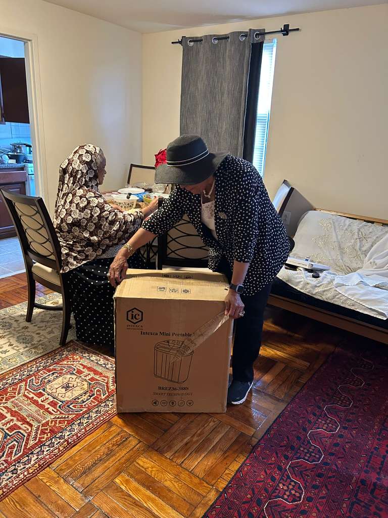 A member kneels to deliver a boxed portable washing machine to an older woman seated at her dining table.