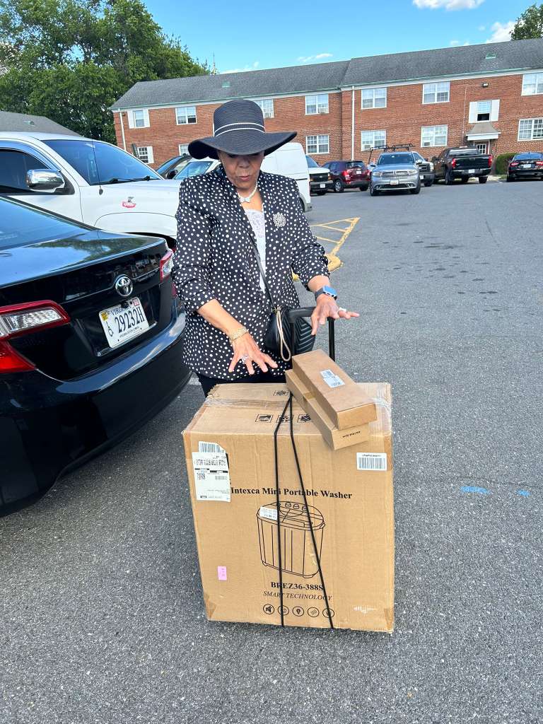 A member loads a large boxed appliance beside a car in an apartment parking lot, ready to deliver it.