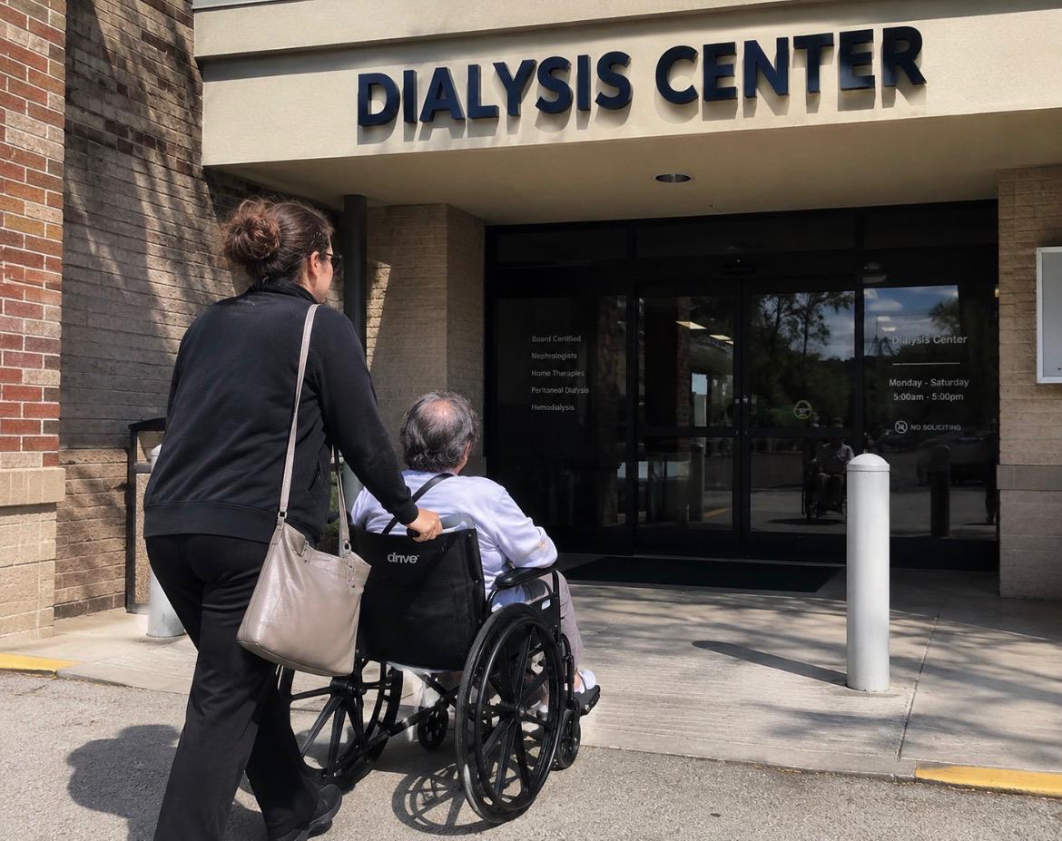 An Alliance member pushes a patient in a wheelchair toward the entrance of a dialysis center on a sunny morning.